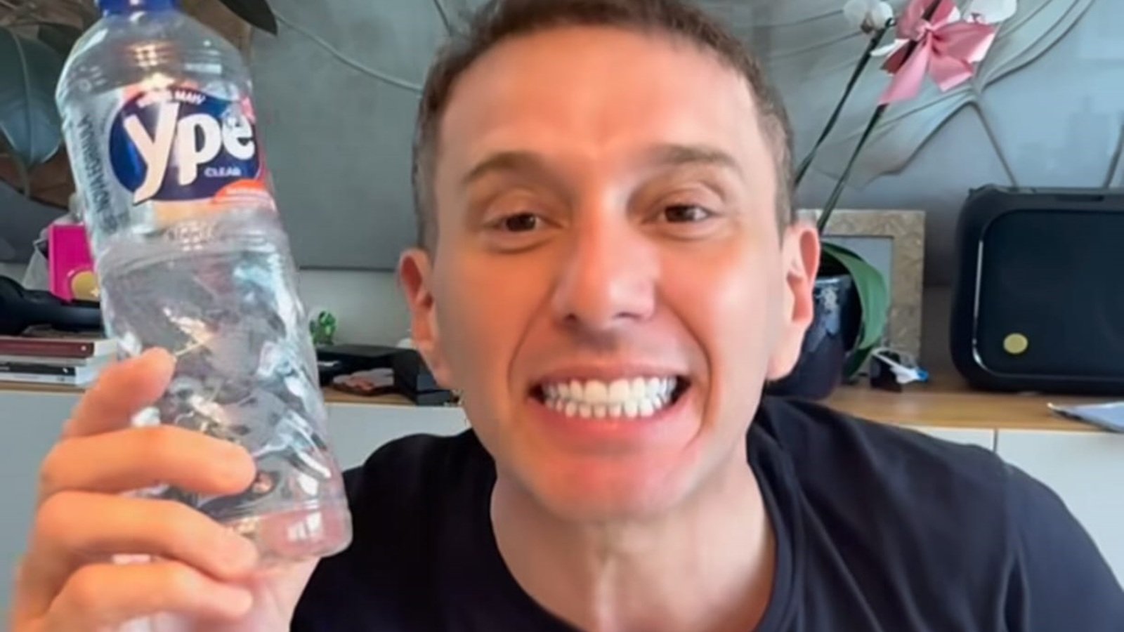 Smiling man in a black shirt holds up a clear plastic water bottle toward the camera blue label visible on the bottle
