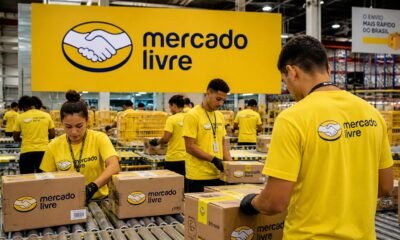 Warehouse workers in yellow Mercado Livre shirts seal and label cardboard boxes on a packaging line in a busy fulfillment center