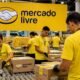 Warehouse workers in yellow Mercado Livre shirts seal and label cardboard boxes on a packaging line in a busy fulfillment center