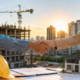 Handshake between a construction worker in an orange safety vest and a businesswoman at a building site with a model house and blueprints on the table
