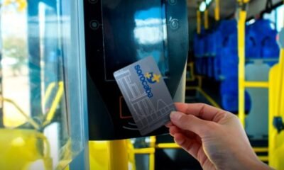 Close-up of a hand holding a contactless transit card near a bus fare reader inside a bus, with blue seats and yellow poles in view.