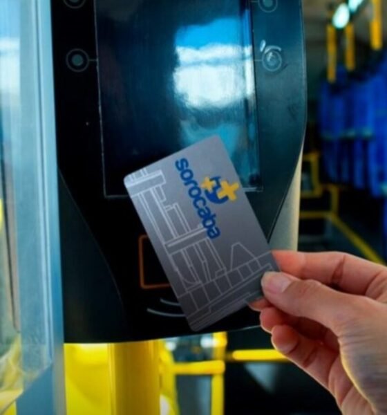 Close up of a hand holding a contactless transit card near a bus fare reader inside a bus with blue seats and yellow poles in view