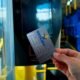 Close up of a hand holding a contactless transit card near a bus fare reader inside a bus with blue seats and yellow poles in view