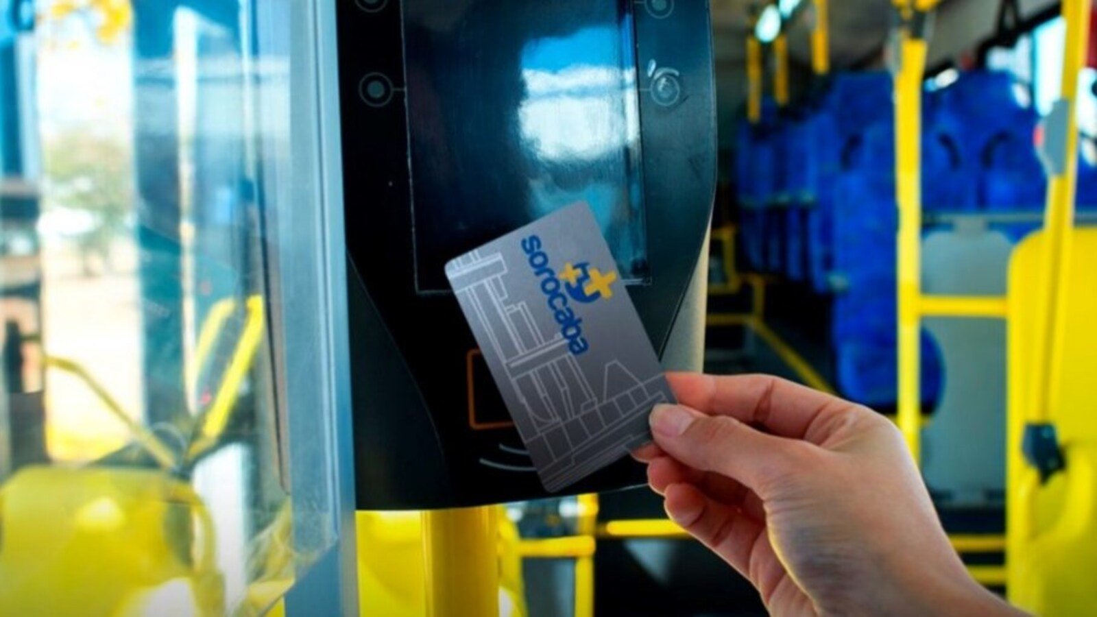 Close up of a hand holding a contactless transit card near a bus fare reader inside a bus with blue seats and yellow poles in view