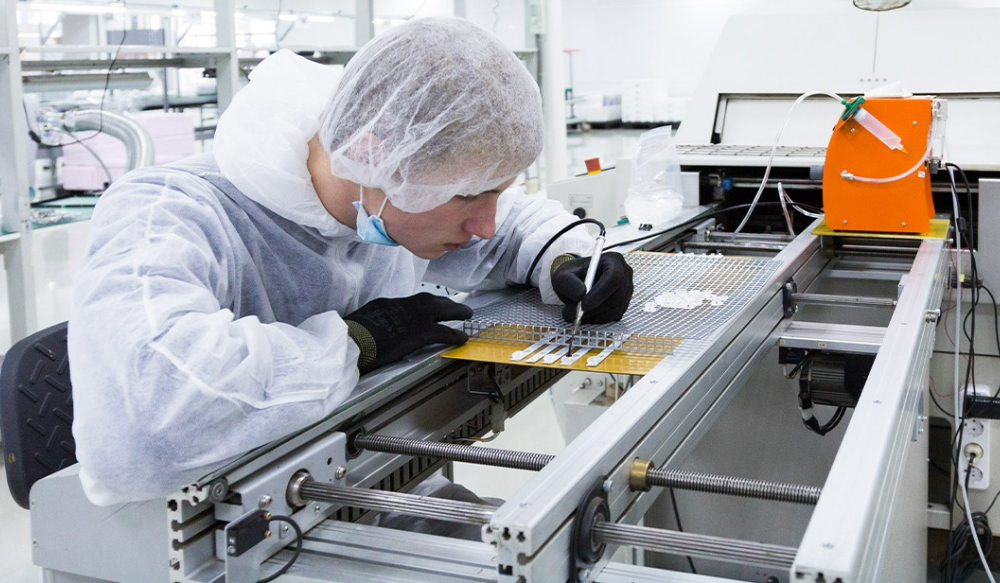 Lab technician in a cleanroom suit and hairnet inspects a PCB on a production line with a precision tool nearby.
