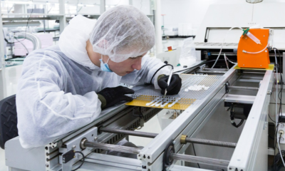 Lab technician in a cleanroom suit and hairnet inspects a PCB on a production line with a precision tool nearby