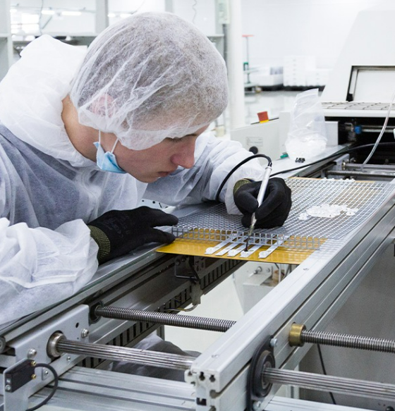 Lab technician in a cleanroom suit and hairnet inspects a PCB on a production line with a precision tool nearby