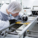 Lab technician in a cleanroom suit and hairnet inspects a PCB on a production line with a precision tool nearby