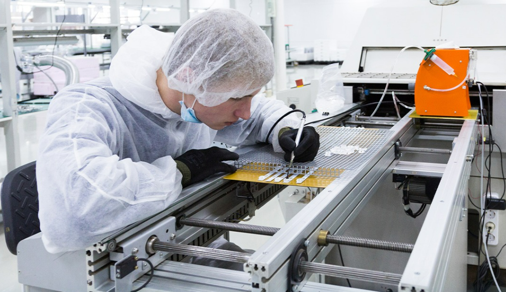 Lab technician in a cleanroom suit and hairnet inspects a PCB on a production line with a precision tool nearby