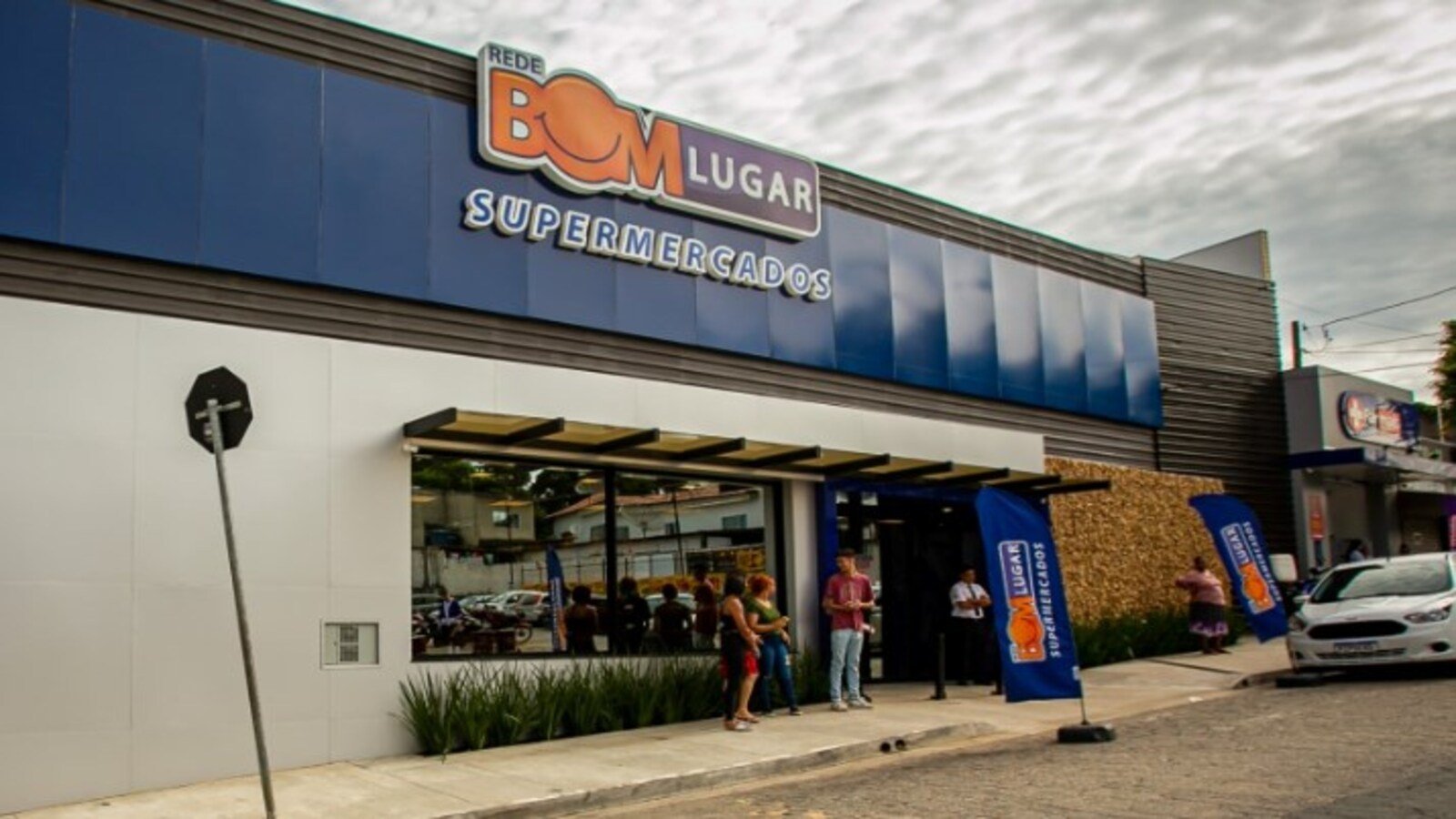 Exterior of Bom Lugar Supermercados storefront with blue panels and glass entrance people standing outside under an awning