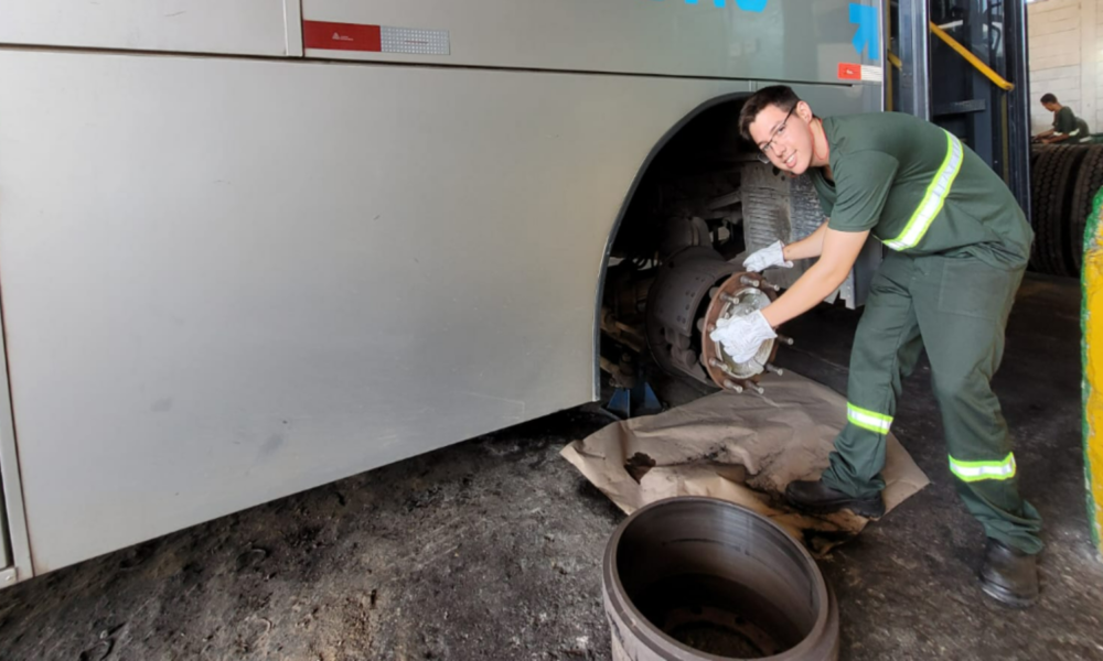 Male mechanic in green coveralls and gloves installing or inspecting a bus wheel brake rotor beside an open wheel in a garage workshop