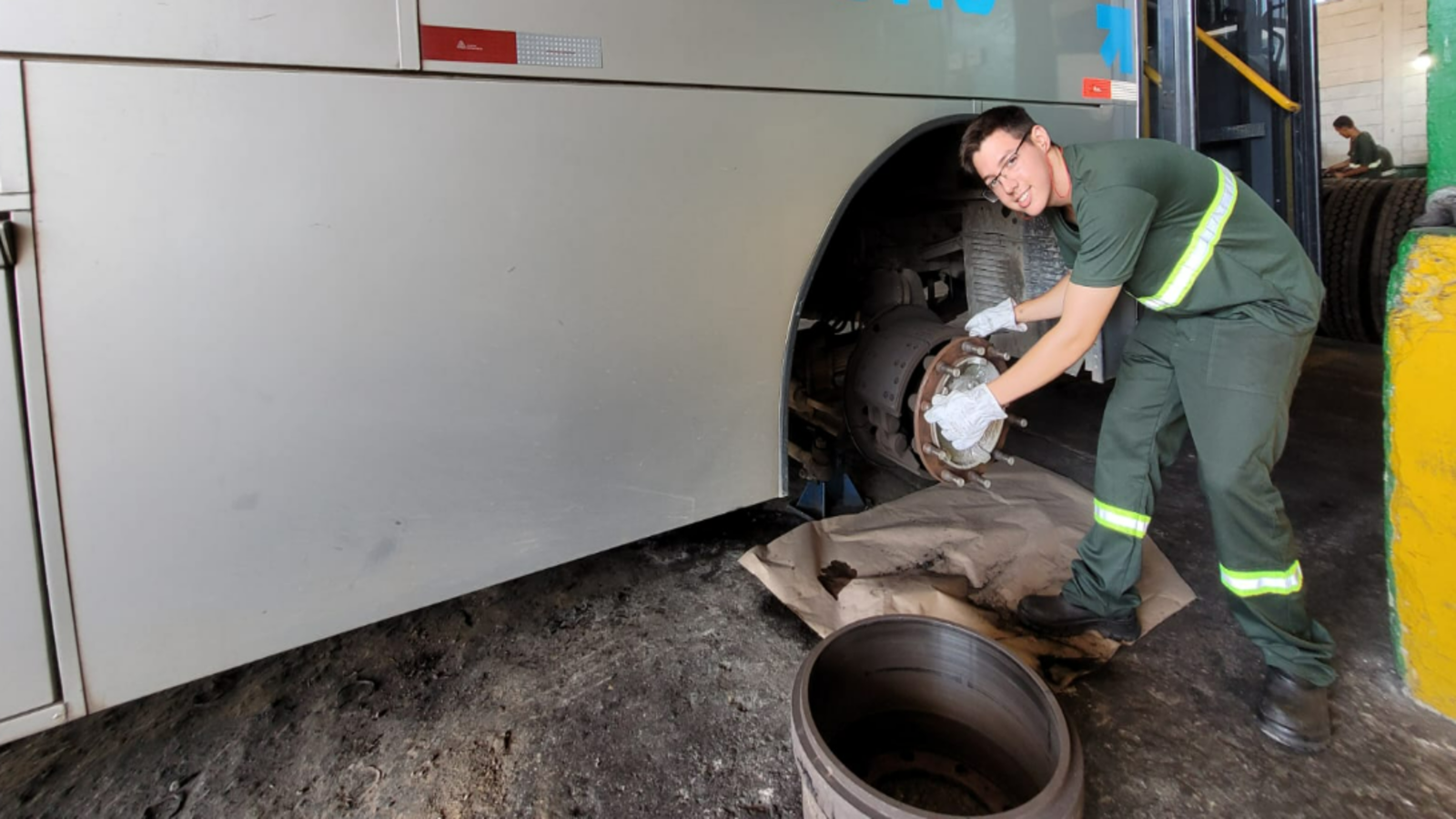 Male mechanic in green coveralls and gloves installing or inspecting a bus wheel brake rotor beside an open wheel in a garage workshop