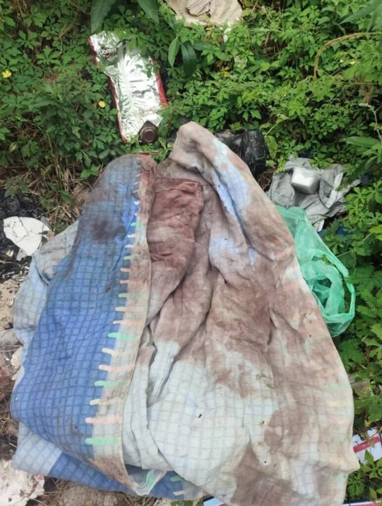 Pile of dirty blankets and clothes discarded in an overgrown outdoor area with litter and a plastic bag nearby