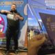 Man posing with a glass trophy in front of a blue event banner that reads Concurso Jornalístico e Publicitário with a large colorful logo and pigeons left panel of a split photo
