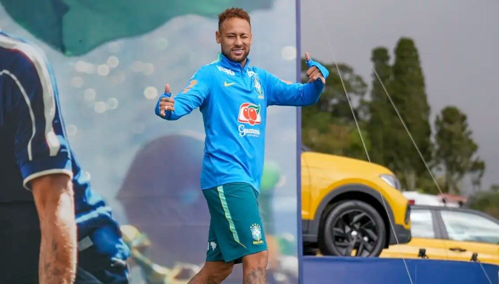 Brazilian footballer in blue training kit giving thumbs up on a sports stage/field, smiling at the camera.