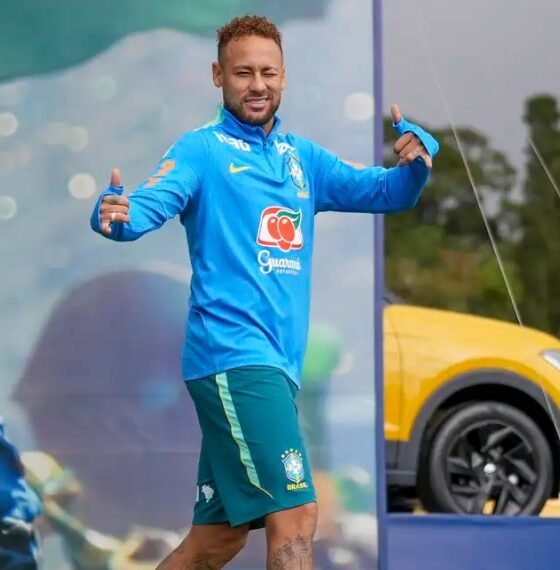 Brazilian footballer in blue training kit giving thumbs up on a sports stage/field, smiling at the camera.