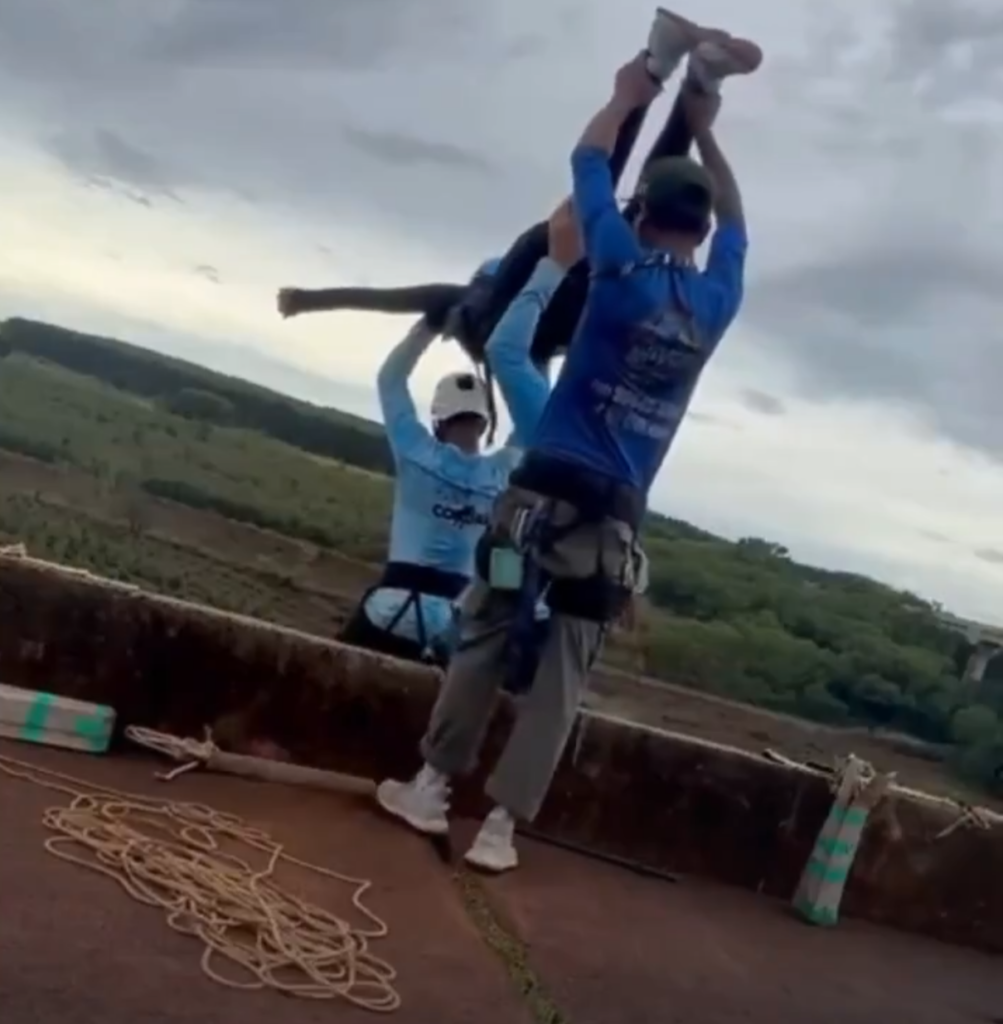 Screenshot_20260613 172518_whatsapp sorocabanices Two rescuers in blue shirts helmets and harnesses lift a teammate overhead with a rope on a concrete platform against a cloudy sky