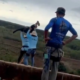Two people in climbing harnesses on a ledge one raising arms as if throwing a rope with a countryside landscape in the background