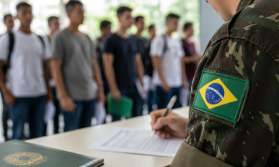 Soldier in camouflage with a Brazilian flag patch signs forms at a desk as recruits line up in the background