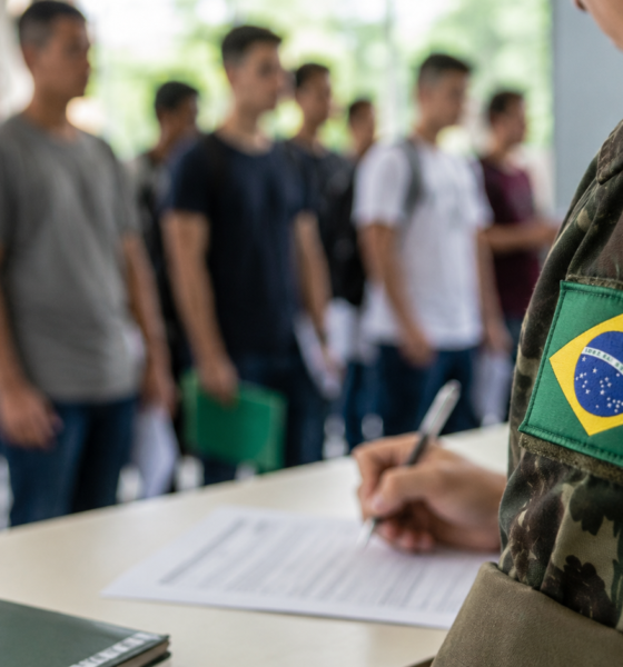 Soldier in camouflage with a Brazilian flag patch signs forms at a desk as recruits line up in the background