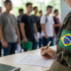 Soldier in camouflage with a Brazilian flag patch signs forms at a desk as recruits line up in the background