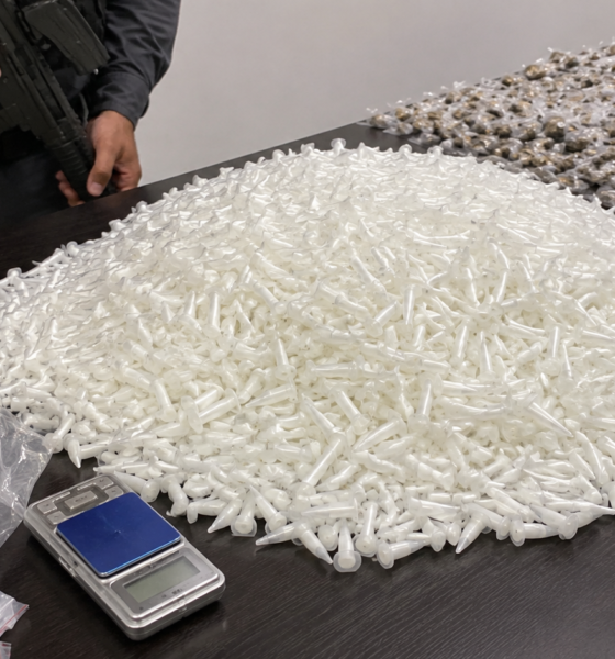 Pile of white plastic syringe tips spread across a table with a police officer and a firearm visible nearby plus a scale and evidence bags in the foreground