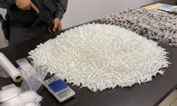 Pile of white plastic syringe tips spread across a table with a police officer and a firearm visible nearby plus a scale and evidence bags in the foreground