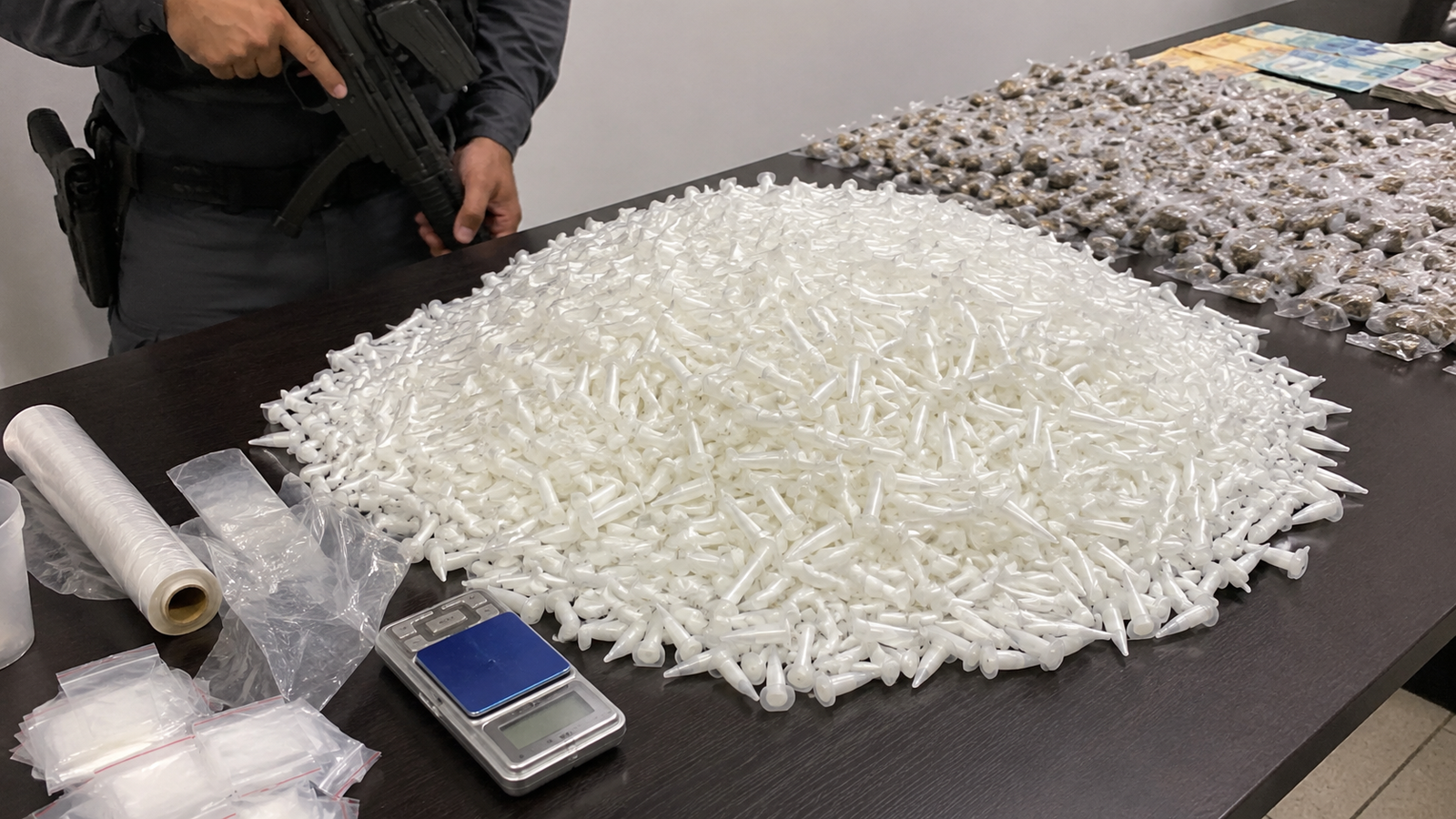 Pile of white plastic syringe tips spread across a table with a police officer and a firearm visible nearby plus a scale and evidence bags in the foreground