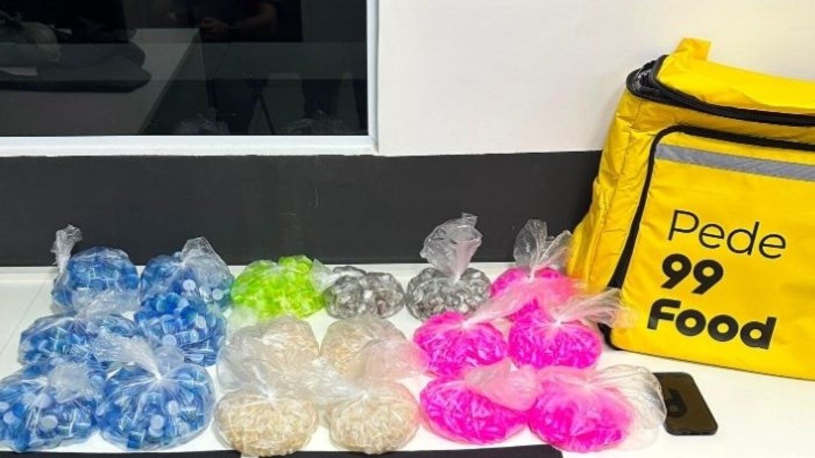 Colorful bags of small snacks in plastic wrap arranged on a white counter beside a yellow delivery bag labeled Pede  Food and a phone