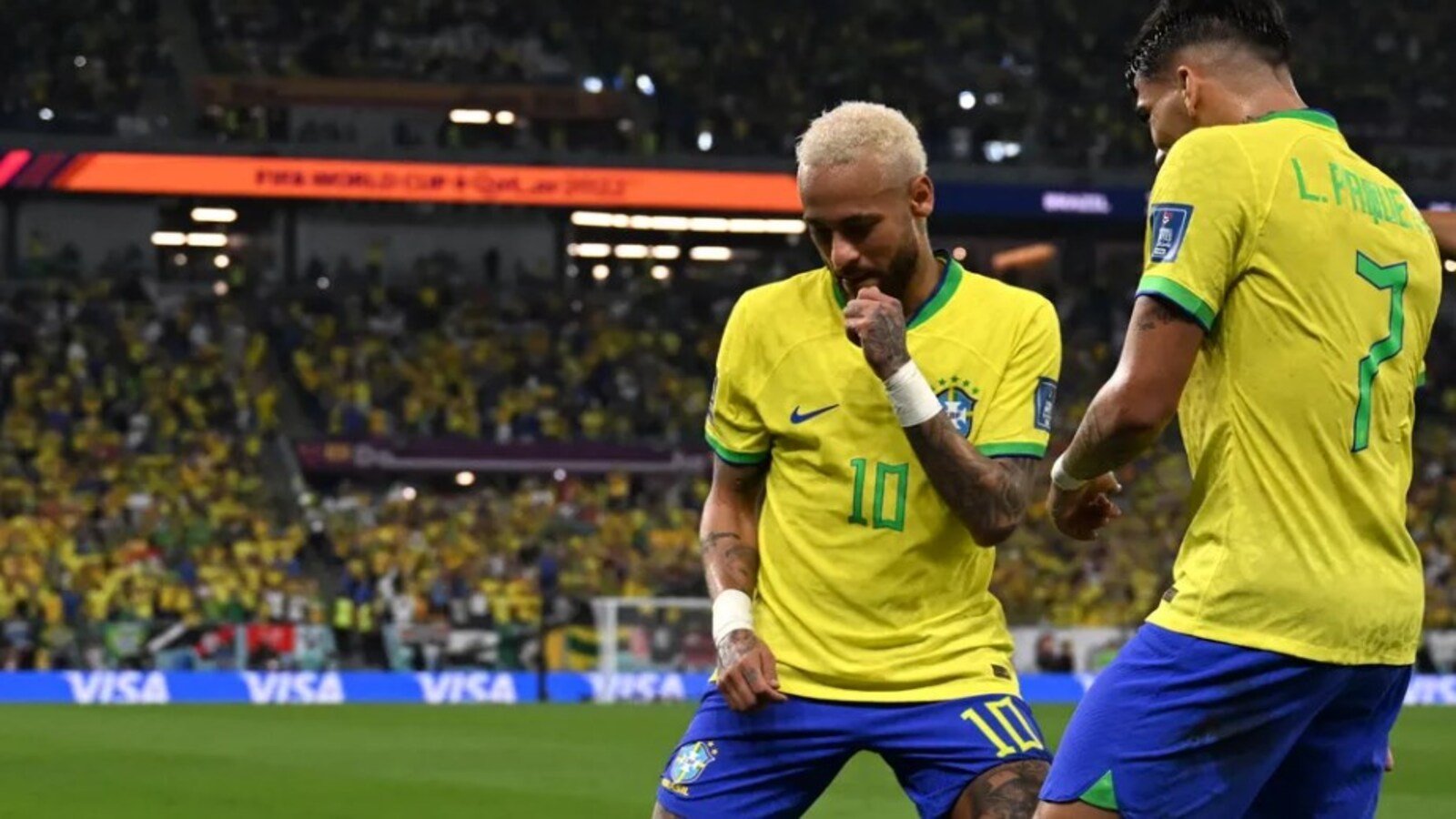 Two Brazil players in yellow jerseys celebrate on the pitch as a full stadium looks on