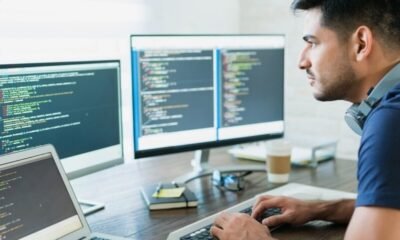 Male developer typing code at a desk with three monitors displaying code and a laptop nearby.