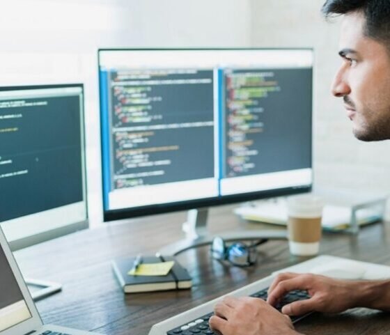 Male developer typing code at a desk with three monitors displaying code and a laptop nearby