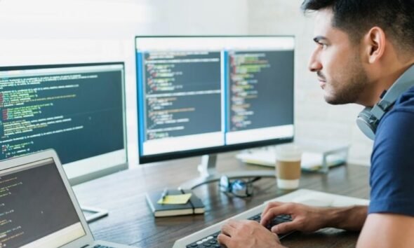 Male developer typing code at a desk with three monitors displaying code and a laptop nearby