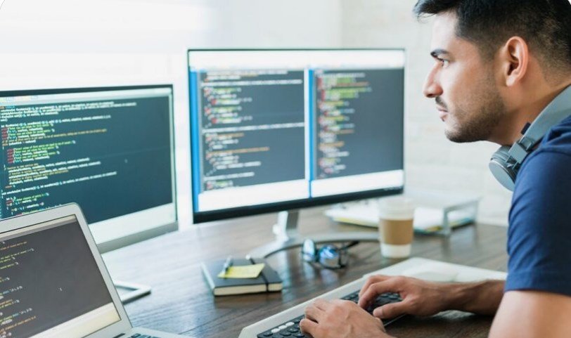 Male developer typing code at a desk with three monitors displaying code and a laptop nearby