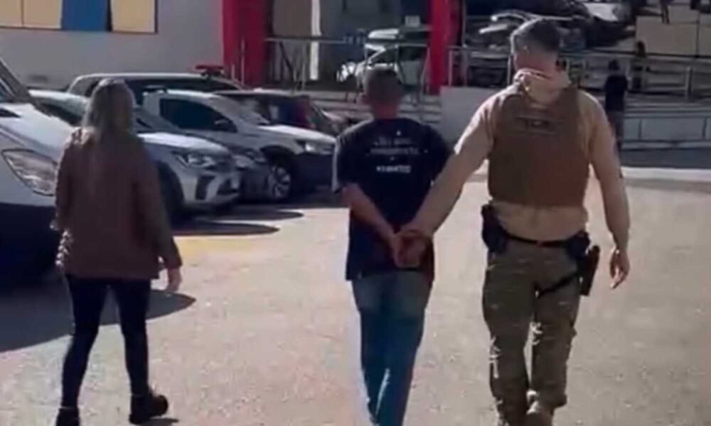 A uniformed officer in tan gear escorts a handcuffed man down a parking lot while a woman walks nearby, with parked cars in the background.
