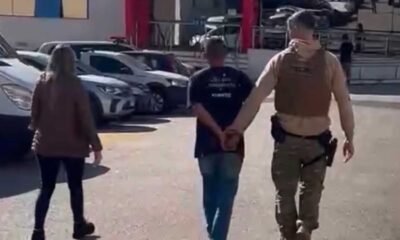 A uniformed officer in tan gear escorts a handcuffed man down a parking lot while a woman walks nearby with parked cars in the background