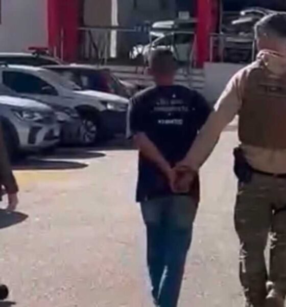 A uniformed officer in tan gear escorts a handcuffed man down a parking lot while a woman walks nearby with parked cars in the background
