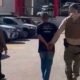 A uniformed officer in tan gear escorts a handcuffed man down a parking lot while a woman walks nearby with parked cars in the background