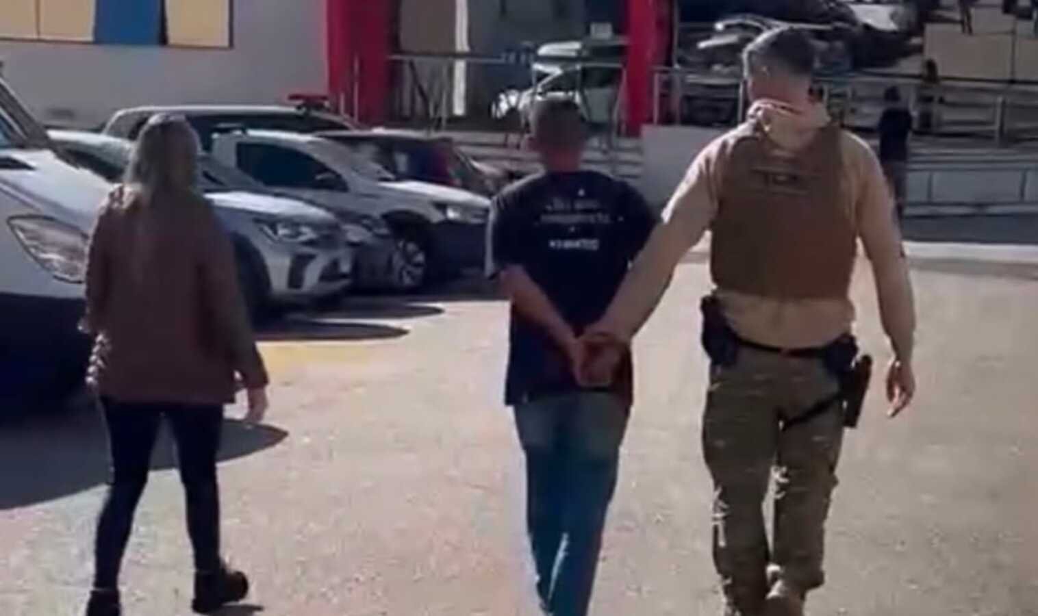 A uniformed officer in tan gear escorts a handcuffed man down a parking lot while a woman walks nearby with parked cars in the background