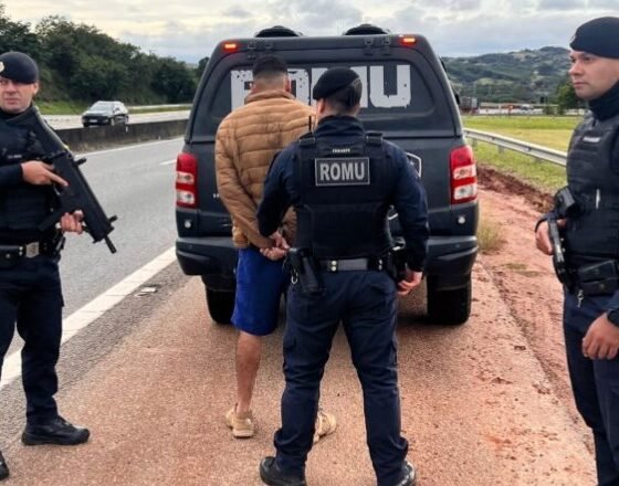 Three police officers detain a man in a tan jacket beside a ROMU marked van on a highway shoulder