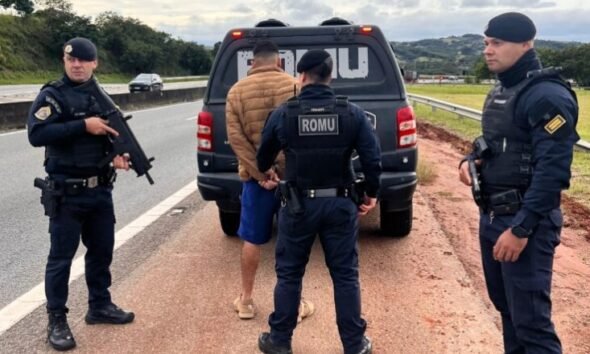 Three police officers detain a man in a tan jacket beside a ROMU marked van on a highway shoulder