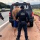 Three police officers detain a man in a tan jacket beside a ROMU marked van on a highway shoulder