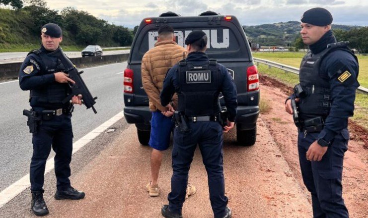 Three police officers detain a man in a tan jacket beside a ROMU marked van on a highway shoulder