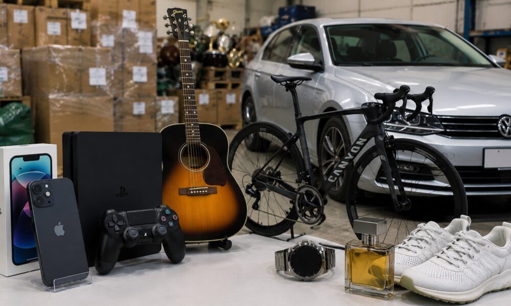 Assorted consumer goods on a table in a warehouse: iPhone box, PS5, guitar, Canyon bike, sneakers, watch, and perfume.
