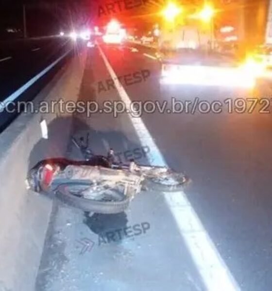 Motorcycle crash on highway at night, lying on its side against a concrete barrier with emergency lights in the distance.