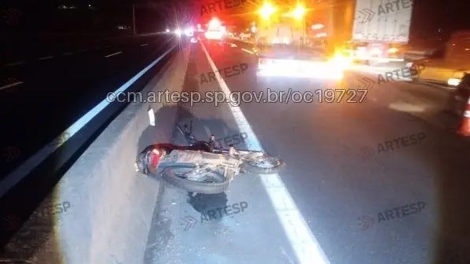 Motorcycle crash on highway at night lying on its side against a concrete barrier with emergency lights in the distance