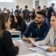 Two recruiters interview a candidate at a crowded career fair with papers and brochures on the table and attendees in the background
