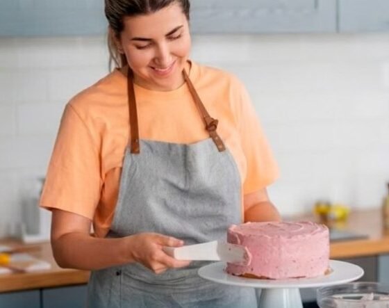 Woman in an orange shirt and gray apron frosting a pink cake on a white cake stand in a bright kitchen