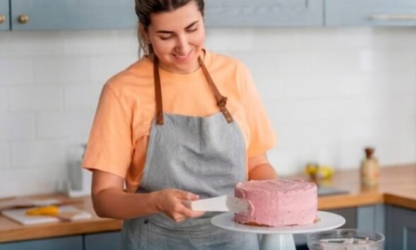Woman in an orange shirt and gray apron frosting a pink cake on a white cake stand in a bright kitchen