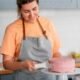 Woman in an orange shirt and gray apron frosting a pink cake on a white cake stand in a bright kitchen